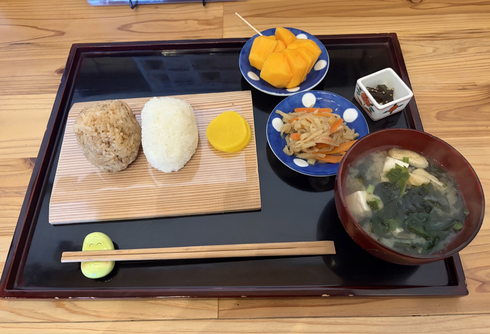 黒い角盆に載ったおにぎり定食。木の板の上に炊き込みご飯と塩のおにぎり、たくあん、かぼちゃの甘煮、切り干し大根、味噌汁