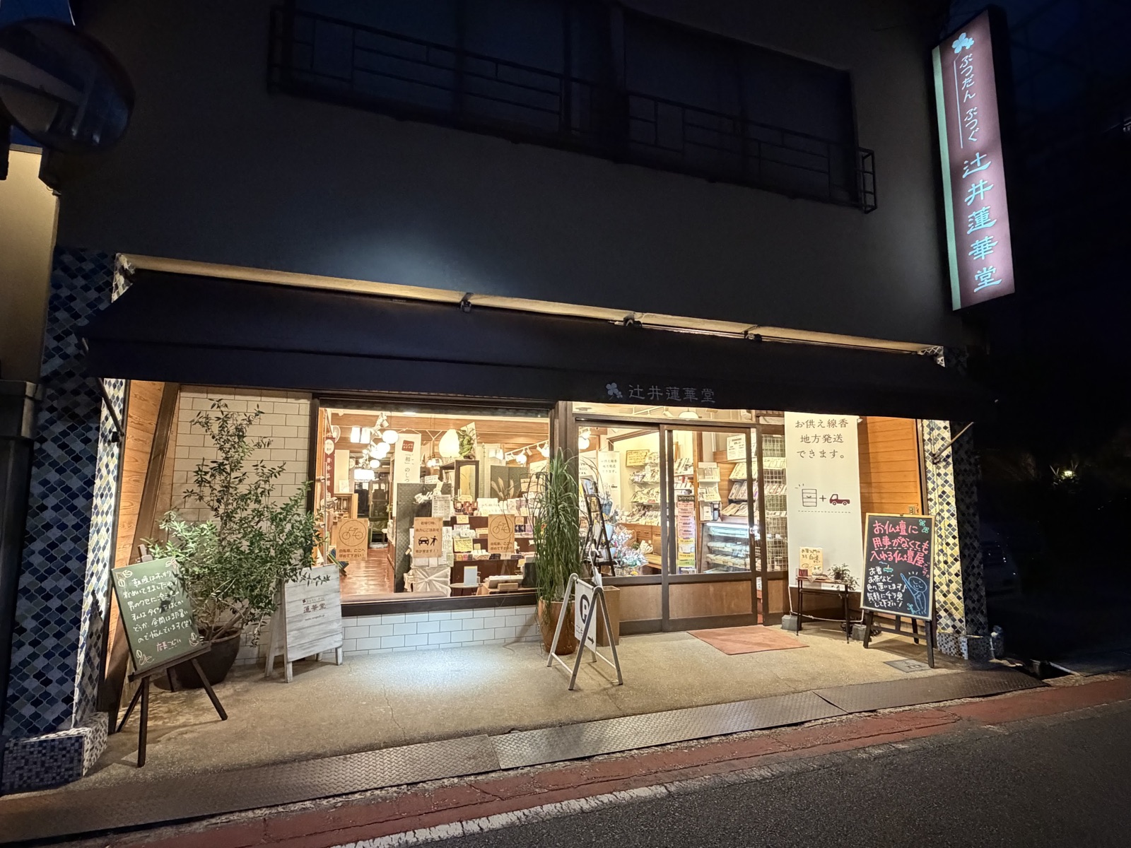 夜の辻井蓮華堂の外観。ピンク色に光る縦看板と、明かりのついたガラス張りの店内。歩道に黒板と鉢植えが並ぶ