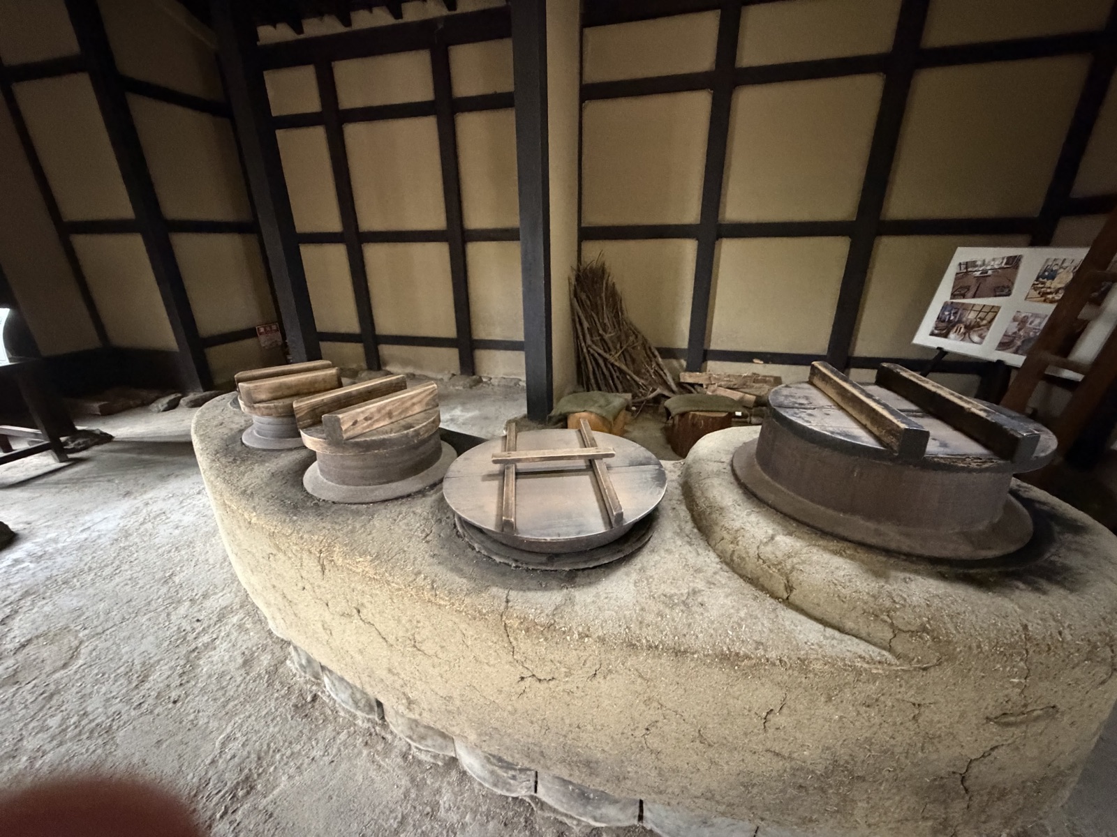 土間に築かれた大きな塗り壁のかまど。丸い木の蓋が4つ並び、奥の壁ぎわに焚き付けの薪が立てかけてある