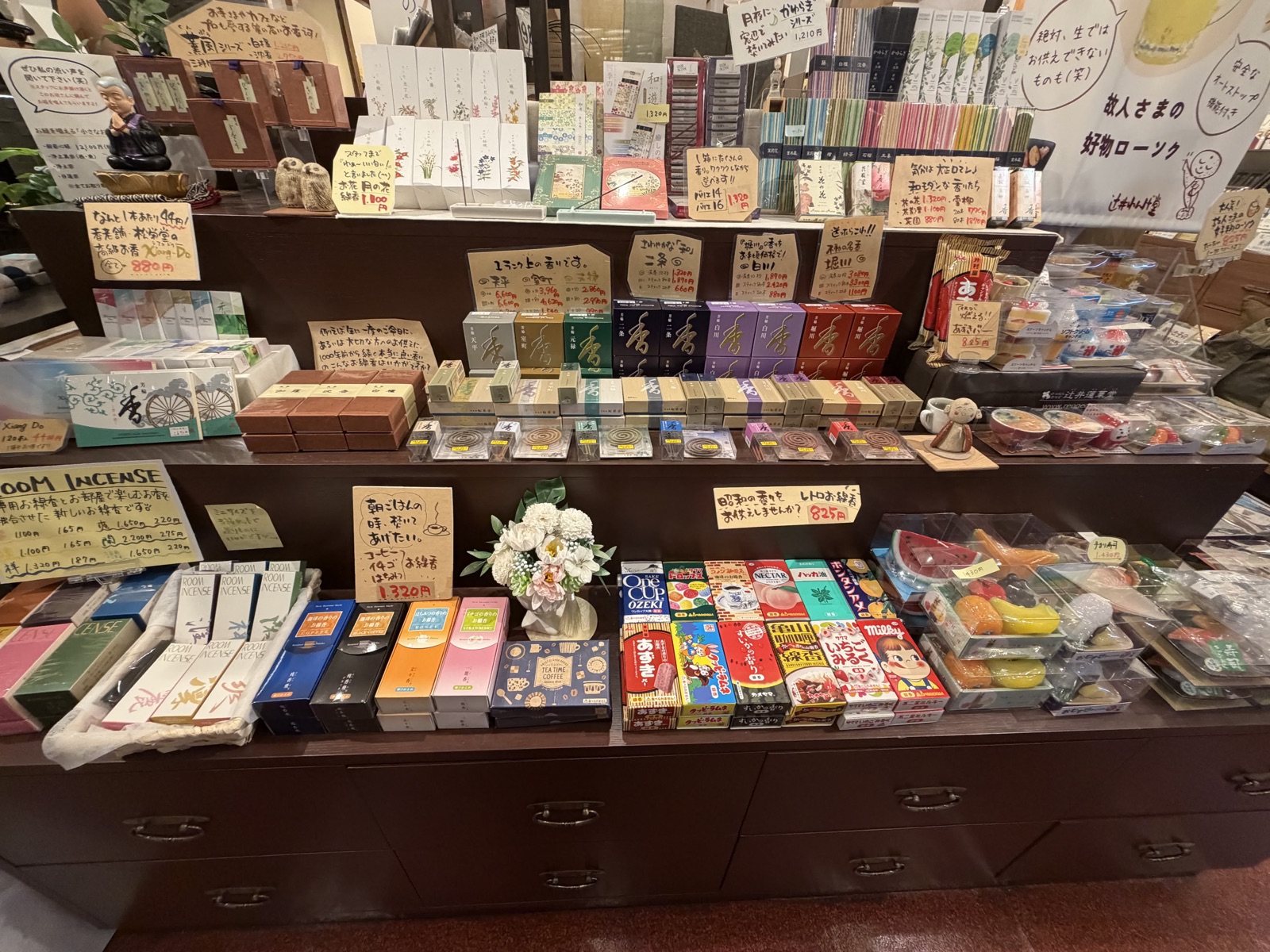 仏具店の棚。お線香の箱が値札とともに並び、右手前にあずきバーやワンカップ、ハッカ飴などのお供え菓子と「昭和の香りをお供えしませんか？ 825円」のPOP