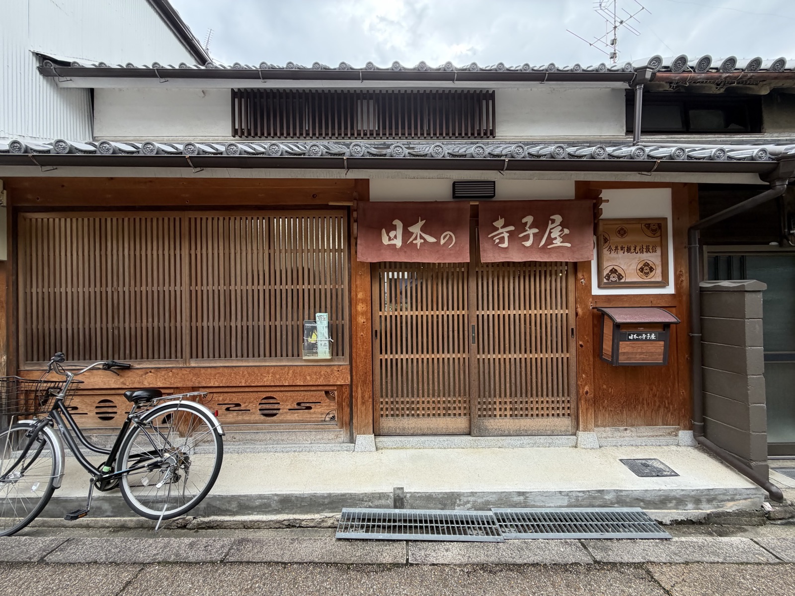 今井町の町家。格子戸と瓦屋根が連なり、「日本の寺子屋」と染め抜いた暖簾。左手に自転車が停めてある