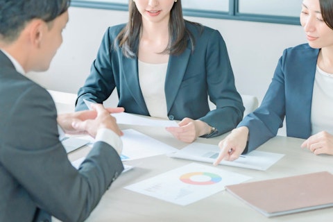 会議室でビジネスウェアを着た3人の男女がテーブルを囲み、円グラフや資料を指差しながら打ち合わせをしている様子