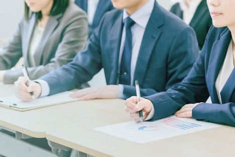 ビジネススーツを着た男女数名が会議室のテーブルに並んで座り、手元の資料やクリップボードにペンで書き込みをしている様子