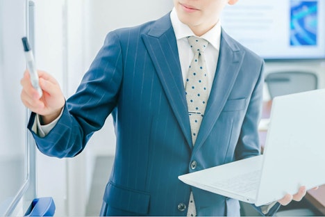 青いストライプのスーツにドット柄のネクタイを締めた男性が、左手に白いノートパソコンを持ちながら、右手のペンでホワイトボードを指し示しているビジネスシーンのクローズアップ画像。