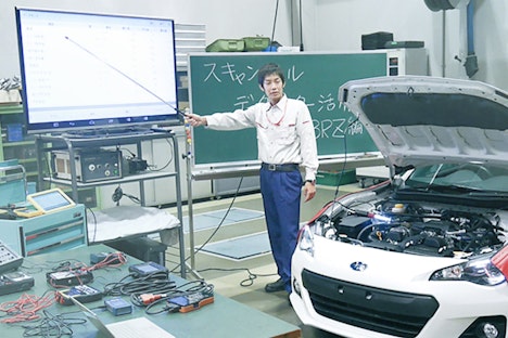 自動車のエンジンルームを開けた白いスポーツカーの横で、作業服を着た男性が指示棒を使いながら大型モニターに映るデータ表を指し示し、整備やデータ活用について解説している研修施設のような室内の様子。