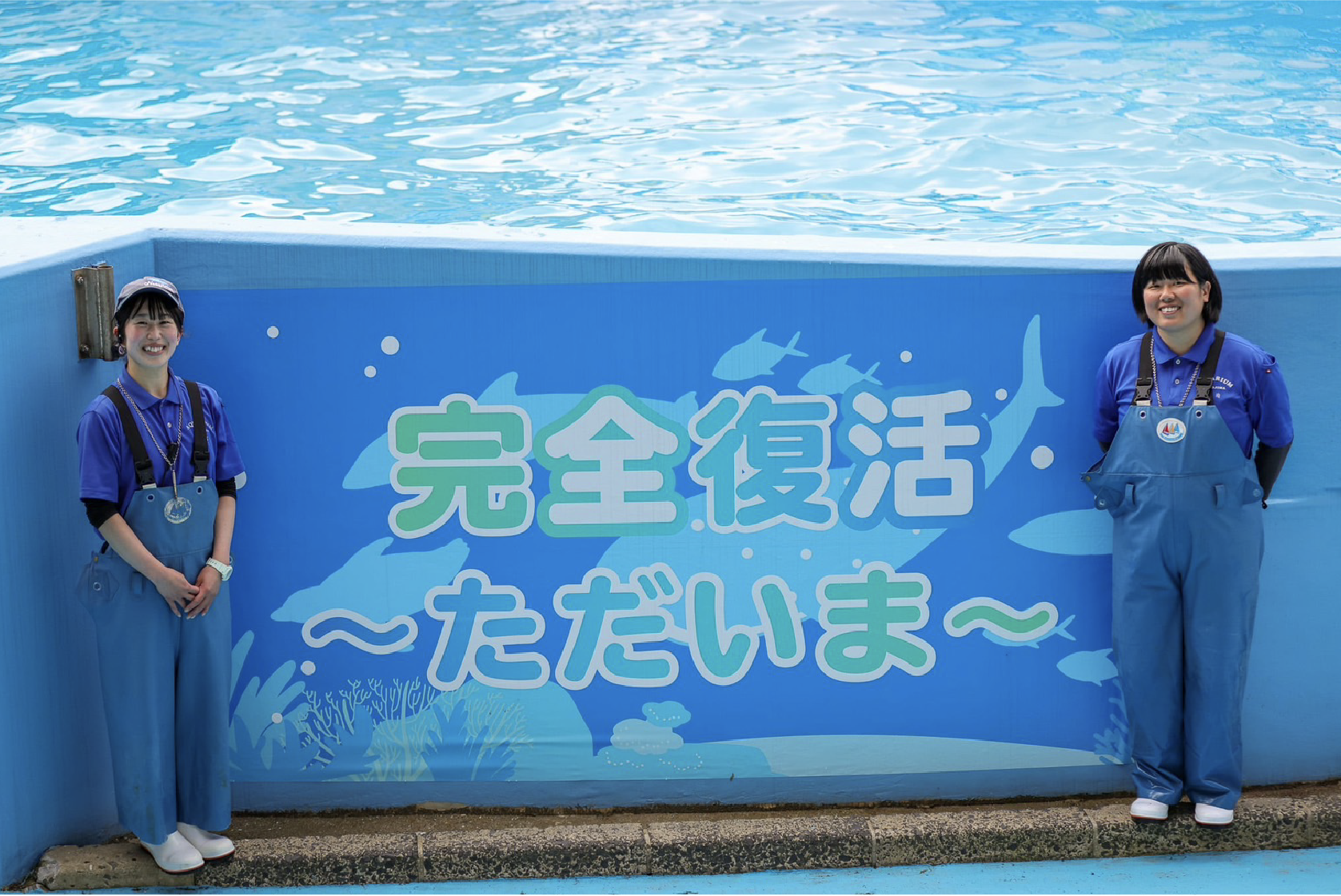 水族館で働く女性スタッフたち。「完全復活～ ただいま～」の横断幕を挟んで左が釘宮ひなたさん、右が 新治奈々花さん。「ぜひ遊びに来て動物たち、水族館の魅力を知ってほしいです！」