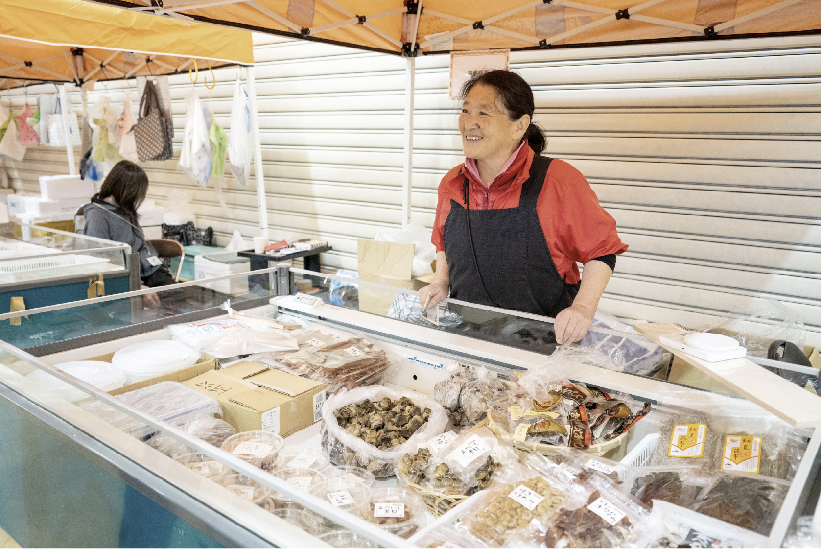 ワイプラザ輪島店のコンコースに並ぶ出張輪島朝市の露店。「今日は電球を忘れてきちゃって。ちょっと暗くてごめんね」。テントの上を指しながら、優しく声を掛けてくれたのは遠島商店の遠島孝子さん。貝やアワビなどの海産物を販売している